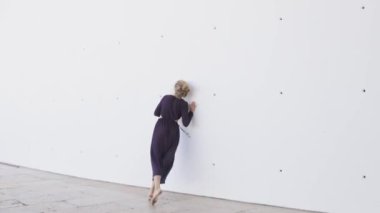 Uzun elbiseli kadın beyaz bir duvarın önünde yalınayak dans ediyor.