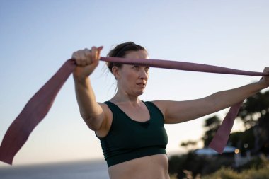 Formda bir kadın dışarıda egzersiz yaparken direniş bandı kullanıyor.