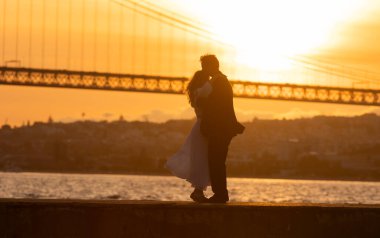 Gün batımında öpüşen bir çiftin silüeti ve arka planda bir köprü ve şehir manzarası.