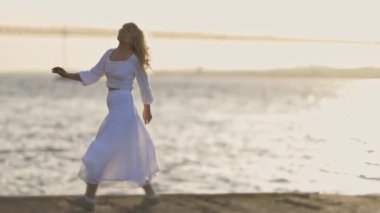 Beyaz elbiseli bir kadın kumsalda zarif bir şekilde dans ediyor. Hareketleri, güneş arka planda batarken denizin ritmiyle akıyor.