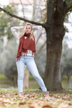 Sonbaharda parkta poz veren, kırmızı atkı takan ve açık mavi kot giyen manken.