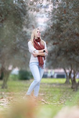 Sarışın bir kadın, zeytin ağaçlarıyla parkta dikilip sonbahar gününün tadını çıkarıyor.