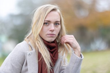 Sonbaharda parkta poz verirken paltosu ve atkısı olan genç sarışın kadın saçına dokunuyor.