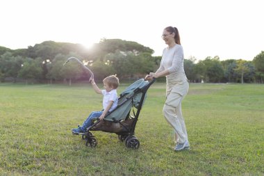 Anne, oğlunu bir oyuncakla oynarken bir parkta bebek arabasına itiyor.