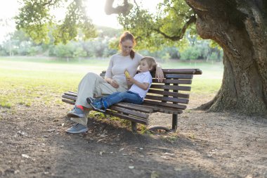 Anne ve oğlu, batan güneşin sıcacık parıltısıyla parktaki bankta huzurlu bir anın tadını çıkarıyorlar.