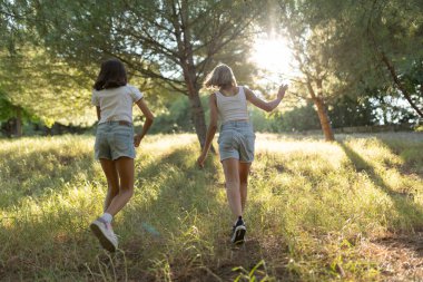 Gün batımında çayırda yürüyen iki kız ağaçların arasından süzülen güneş ışığının tadını çıkarıyorlar.