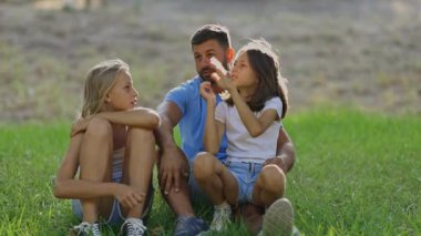 Baba çimlerin üzerinde oturan iki üzgün kızını teselli ediyor.