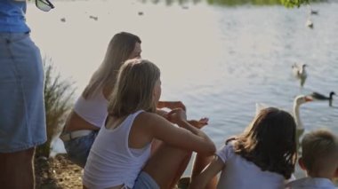 Aile güneşli bir günün tadını çıkarıyor, göl kenarında ördek ve kaz besliyor.