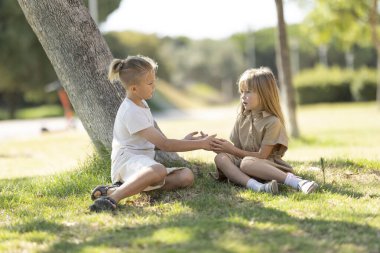 İki çocuk çimenlerde oturuyor, güneşli bir parkta bir ağacın altında konuşuyor.
