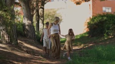 Sıcak bir öğleden sonra sahnesinde bir anne, iki çocuğuyla birlikte yeşil ve ağaçlarla çevrili, güneşli, sakin bir yolda yürüyor.
