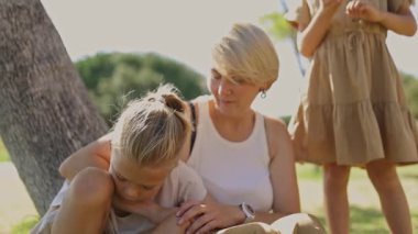 Aile parkta güneşli bir günün tadını çıkarır, bir ağacın altında oturup, bir annenin sevgisini ve çocuklarının neşesini gösterir.