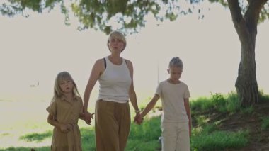 Anne ve iki çocuk, güneşli bir günde parkın içinde el ele tutuşup doğanın tadını çıkarıyorlar.