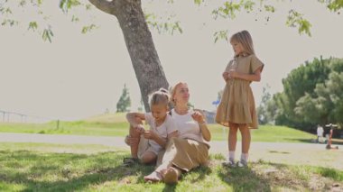 Çocuklar güneşli bir park ortamında bir ağacın gölgesinde dinleniyor ve birbirleriyle etkileşiyorlar.
