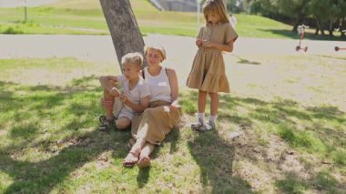 Anne ve çocuklar güneşli bir parkta bir ağacın altında rahatlıyor, birbirlerine eşlik ediyorlar. Bir anlık aile bağı ve doğa takdiri.