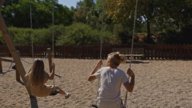 Çocuklar güneşli bir parkta sallanırken keyif alırlar, yemyeşil ve açık gökyüzünün ortasında neşeli anların tadını çıkarırlar.