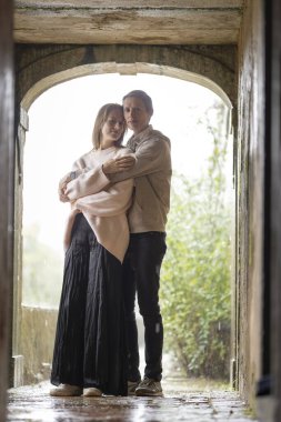 Dışarıda yağmur yağarken taştan bir kemerde sarılan çift romantik ve samimi bir sahne yaratıyor.