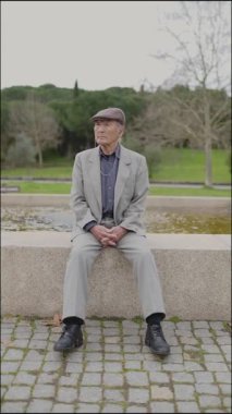 Takım elbiseli ve düz şapkalı yaşlı bir adam parkta alçak bir duvarda oturur ve çevreyi izler.