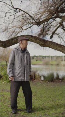 Kıdemli adam gölün kenarındaki bir ağacın altında huzur içinde duruyor, parkın huzurunun tadını çıkarıyor.