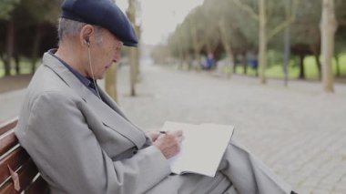 Kulaklık takan son sınıf öğrencisi bankta otururken deftere yazıyor.