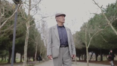 Takım elbiseli ve şapkalı bir adam parkta duruyor. Bir koşucu geçerken düşüncelere dalmış gibi görünüyor.