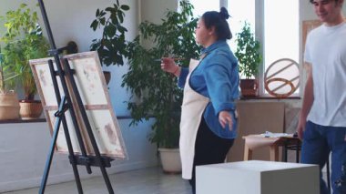 Bir kadın ressam tuvale resim çiziyor referans olarak bir erkek mankeni alıyor - erkek kadının önüne oturuyor. Orta çekim