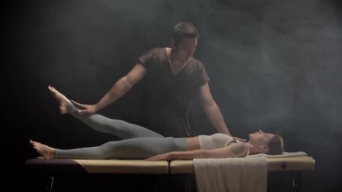 Genç bir kadın, karanlık dumanlı bir odada masaj masasında bir fizyoterapistten bacak gerdirme ameliyatı oluyor.