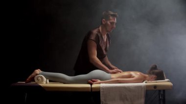 Profesyonel masör, karanlık odada dumanla masaj masasında uzanan genç bir kadına rahatlatıcı sırt masajı yapıyor.