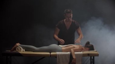 Profesyonel masör, bir kadının vücudunu bir havluyla kaplıyor. Karanlık bir odada, dumanla.