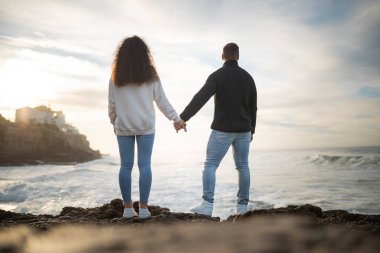 Romantik çift okyanusun üzerinde güzel bir günbatımının tadını çıkarıyor, kayalık bir sahilde el ele duruyorlar.