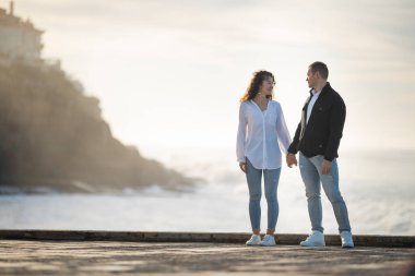 Romantik çift gün batımında deniz kenarında el ele tutuşup birbirlerine bakıyorlar.