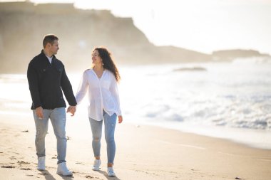 Romantik çift gün batımında kumsalda sakin bir gezinti yapıyor, el ele tutuşuyor ve birlikte hassas bir anı paylaşıyorlar.