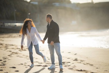 Romantik çift gün batımında kumsalda keyifli bir gezinti yapıyor, el ele tutuşuyor ve neşeli bir anı paylaşıyorlar.