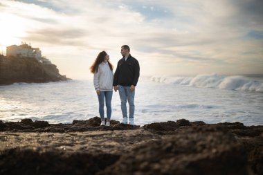 Romantik çift kayalık bir sahilde güzel bir günbatımının tadını çıkarıyor, el ele tutuşuyor ve okyanus dalgalarını seyrediyor.
