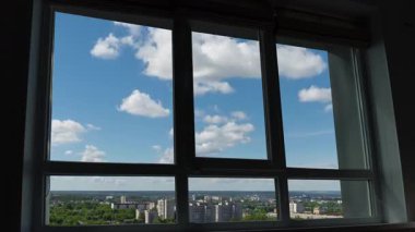 Beyaz bulutlar mavi gökyüzünde sürükleniyor, yüksek katlı bir apartman penceresinden bakılıyor.