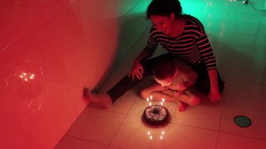 Sevecen bir anne, oğlunun doğum günü pastasına mum üflemesine yardım ediyor. Renkli ışıkların değiştiği bir odada.