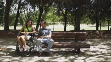 Tekerlekli sandalyede genç bir kadın ve bir adam parkta bir bankta sohbet ediyorlar.