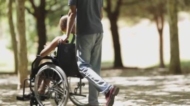 Adam, tekerlekli sandalyedeki genç bir kadını güneşli bir parkta itiyor.
