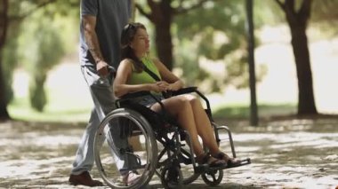 Adam, tekerlekli sandalyedeki bir kadını güneşli bir parkta itiyor.