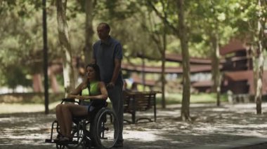 Adam, parkta tekerlekli sandalyedeki genç bir kadını yavaş çekimde itiyor.