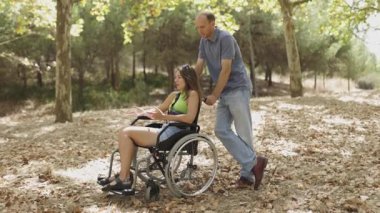 Adam ağır çekimde tekerlekli sandalyede bir kadını ormana itiyor.