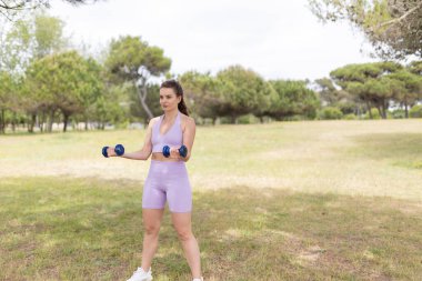 Mor spor kıyafetli genç bir kadın parkta egzersiz yaparken ağırlık kaldırıyor..
