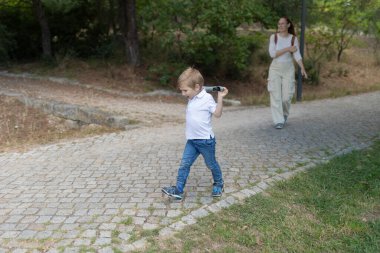 Küçük çocuk annesiyle parkta asfalt bir yolda yürürken dürbünle oynuyor.