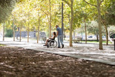 Hemşire, tekerlekli sandalyedeki kadın hastaya güneşli bir parkta yardım ediyor, hareket desteğini sağlıyor ve dışarıda eğleniyor.