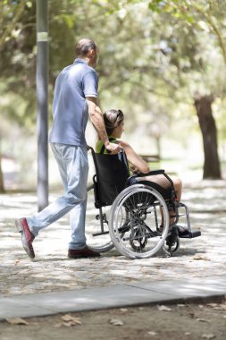 Adam engelli bir kadına tekerlekli sandalyede yardım ediyor, güneşli bir parkta geziniyor.
