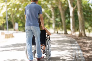 Tekerlekli sandalyedeki engelli kadına yardım eden adam parkta yürüyor.