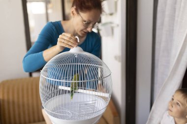 Annesi, içinde yeşil bir papağan olan beyaz, yuvarlak bir kuş kafesini kapatıyor, çocuğu kuşa bakıyor.