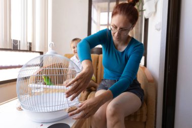 Kafeste muhabbet kuşuyla kuş yemini değiştiren bir kadın, onu izleyen oğlu.