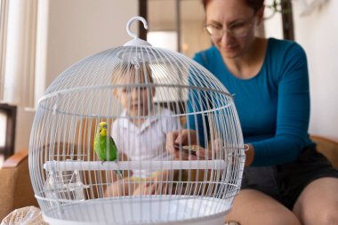 Anne ve çocuk, kafesinin içine tünemiş yeşil bir muhabbet kuşunu besliyor, aile bağlarını ve sorumlu hayvan sahipliğini destekliyor.