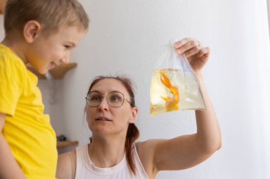 Anne, içinde Japon balığı olan plastik poşeti tutarken gülümseyen çocuğuna gösteriyor.
