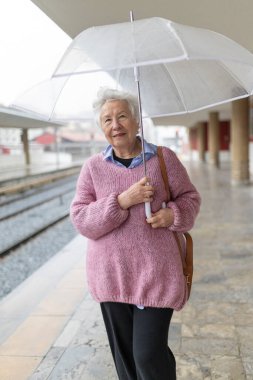 Tren istasyonunda yağmurlu bir günde şeffaf bir şemsiye tutan yaşlı kadın.